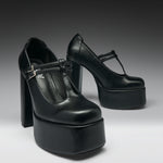 A pair of black leather high-heeled platform Mary Jane shoes with T-strap buckles, photographed on a dark-to-light gray gradient studio background.