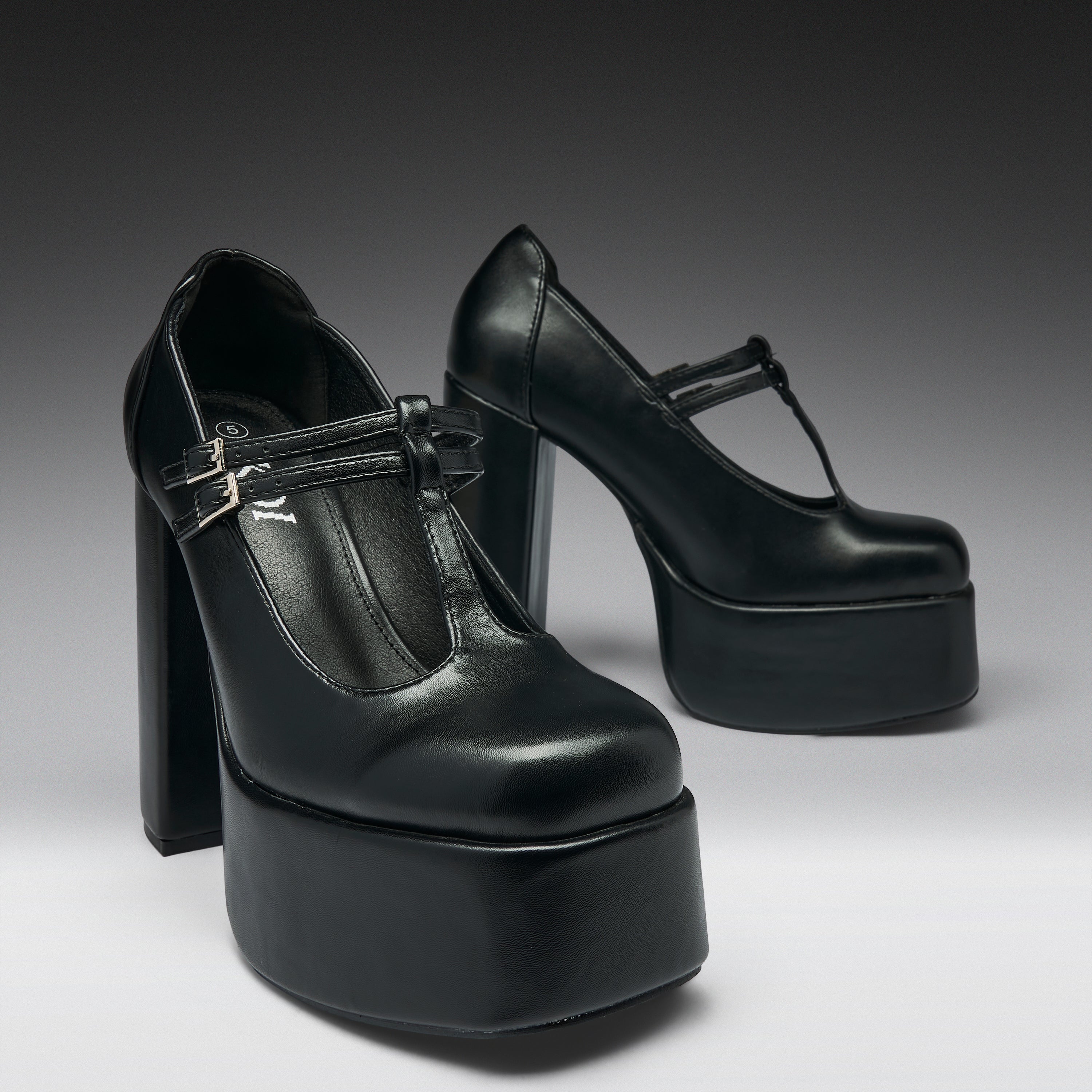 A pair of black leather high-heeled platform Mary Jane shoes with T-strap buckles, photographed on a dark-to-light gray gradient studio background.