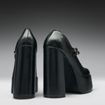 A pair of black leather high-heeled pumps with chunky block heels and platform soles, shown from the back and side against a dark gray gradient background.