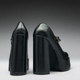 A pair of black leather high-heeled pumps with chunky block heels and platform soles, shown from the back and side against a dark gray gradient background.