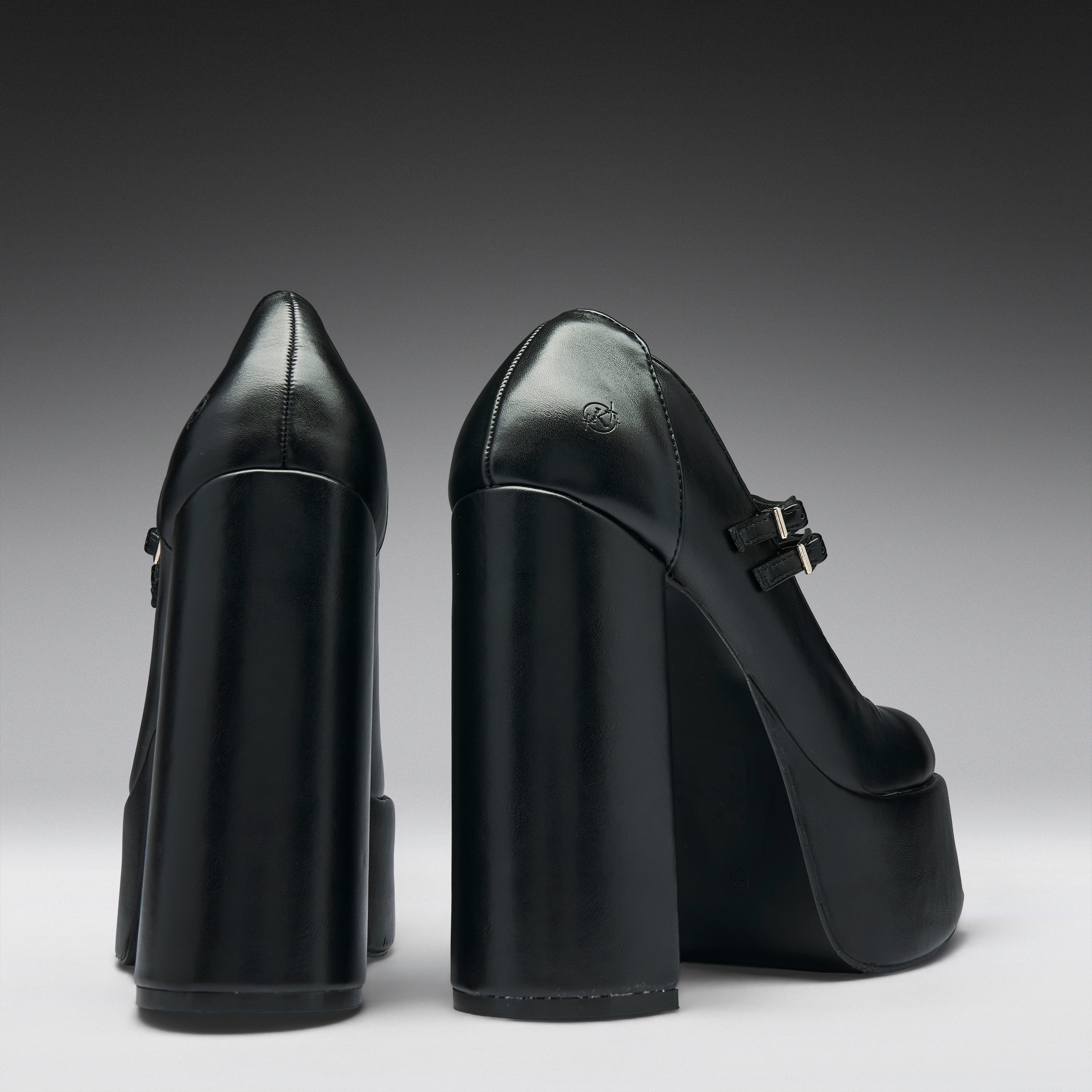 A pair of black leather high-heeled pumps with chunky block heels and platform soles, shown from the back and side against a dark gray gradient background.