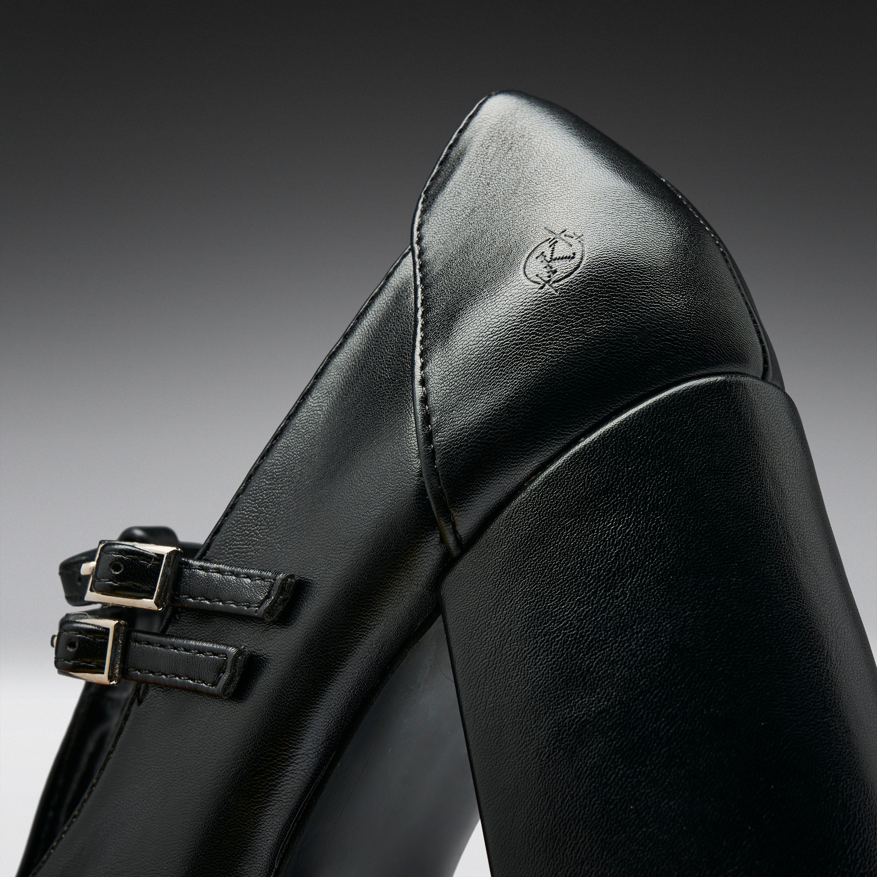 Close-up of a black leather boot with double buckle straps and a small embossed logo on the heel, photographed against a dark gray gradient background.