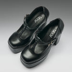 A pair of black leather Mary Jane platform heels with block heels and buckle straps, shown on a neutral gray gradient background. The brand name 'IOKI' is printed on the insoles.