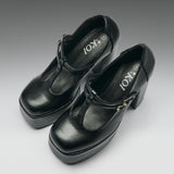 A pair of black leather Mary Jane platform heels with block heels and buckle straps, shown on a neutral gray gradient background. The brand name 'IOKI' is printed on the insoles.