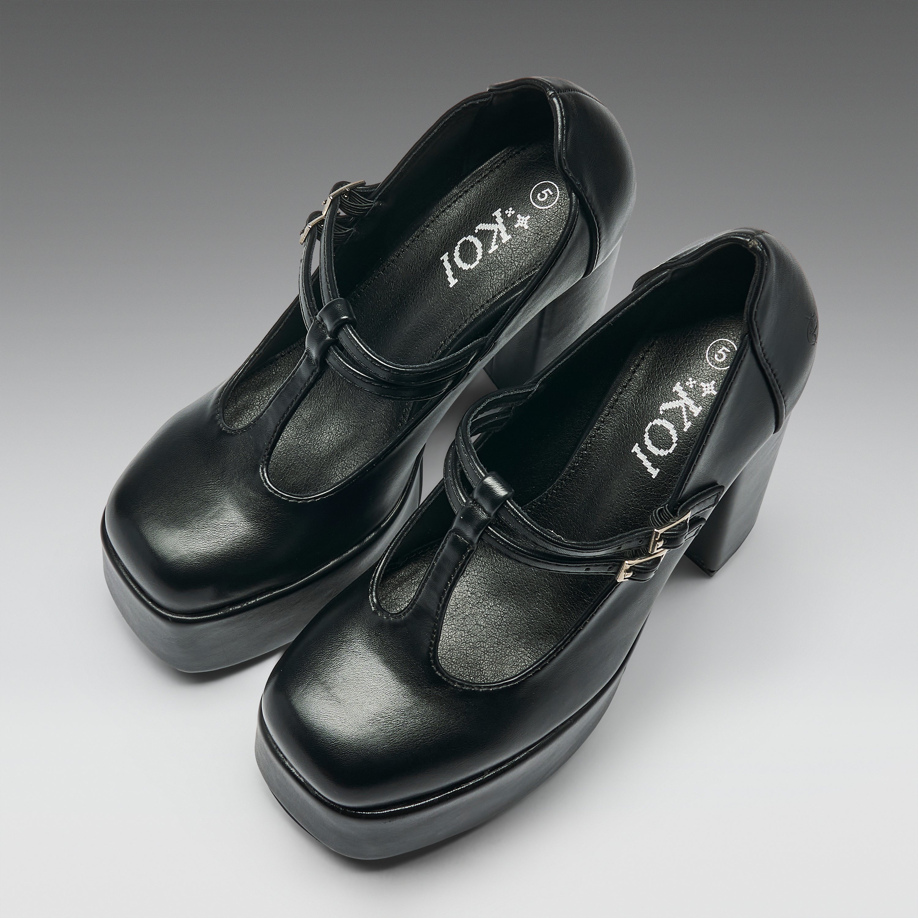 A pair of black leather Mary Jane platform heels with block heels and buckle straps, shown on a neutral gray gradient background. The brand name 'IOKI' is printed on the insoles.
