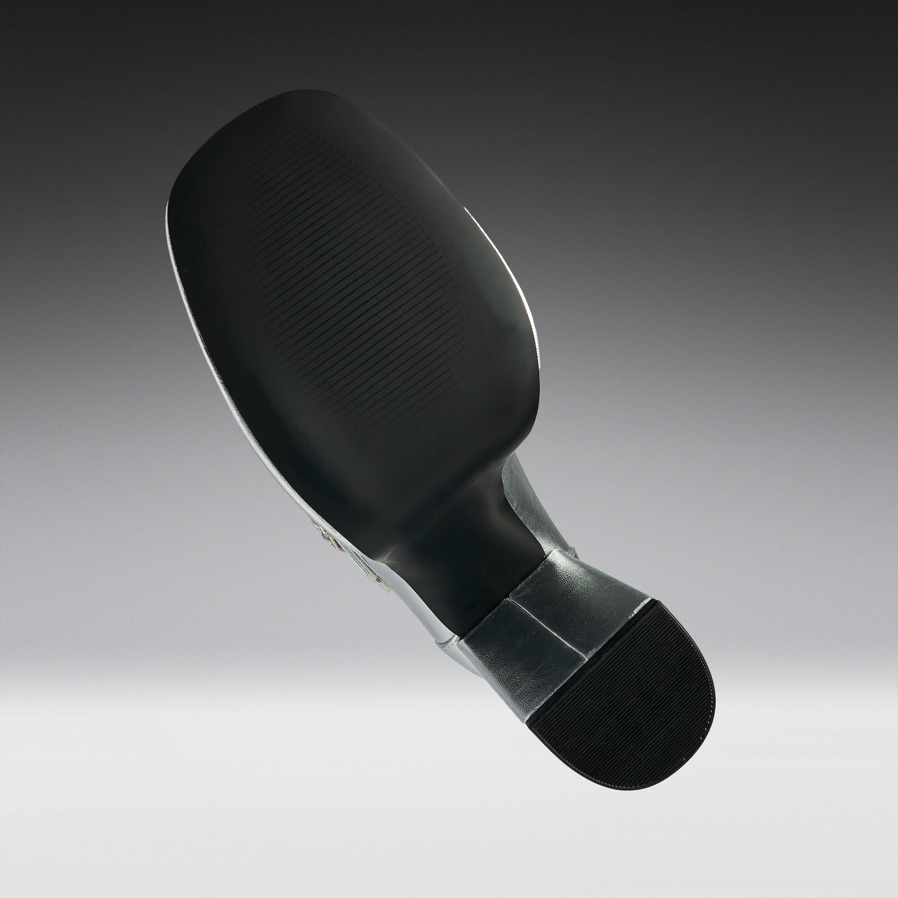A close-up studio shot of the underside of a black athletic shoe, showing the textured outsole, heel, and a small section of the metallic outsole edge against a gray gradient background.