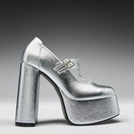 A metallic silver platform high-heeled shoe with a Mary Jane strap and double buckle detail, shown from a side profile on a dark-to-light gray gradient background.