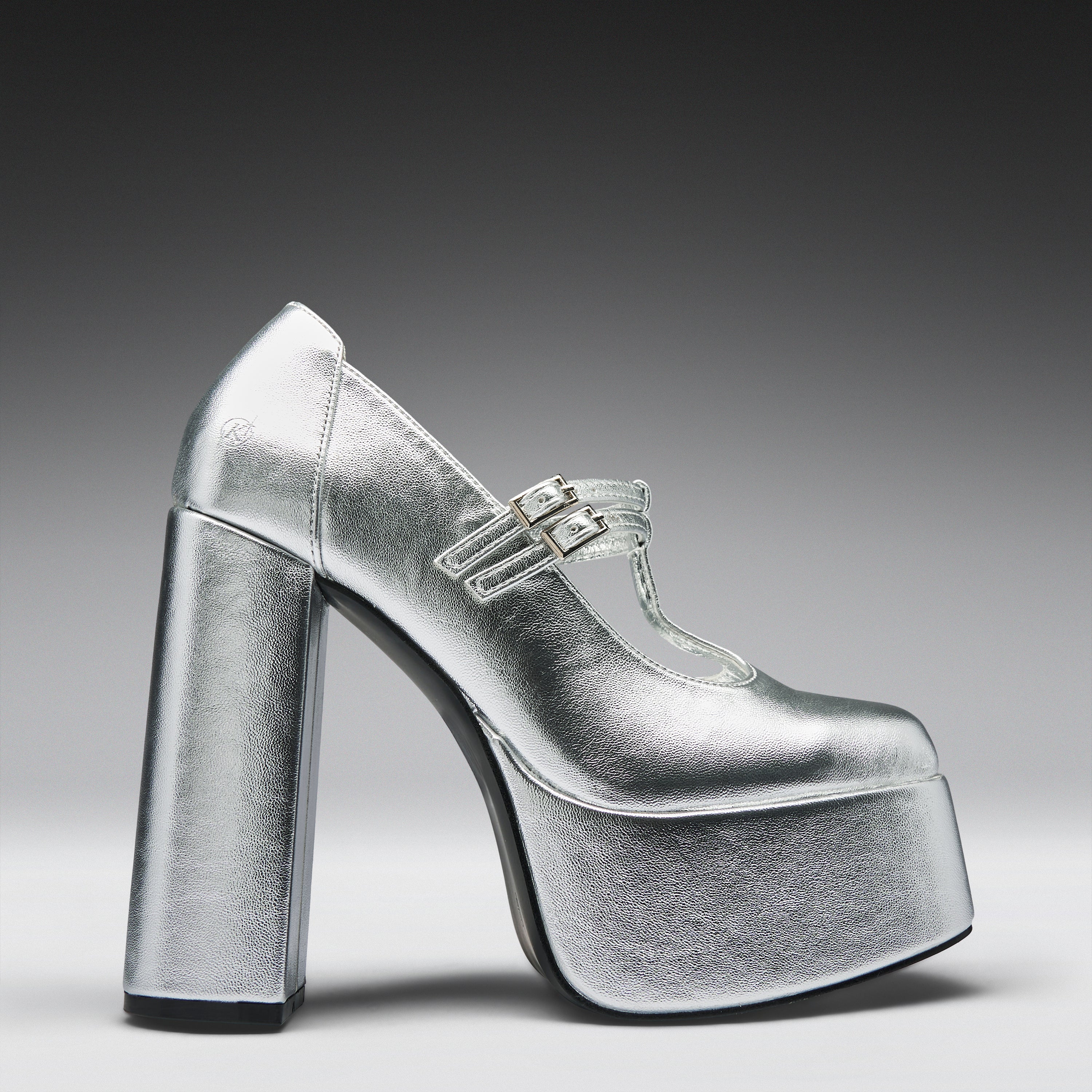A metallic silver platform high-heeled shoe with a Mary Jane strap and double buckle detail, shown from a side profile on a dark-to-light gray gradient background.