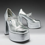 A pair of metallic silver platform Mary Jane high heels with block heels and ankle straps, photographed on a dark-to-light gray gradient studio background.