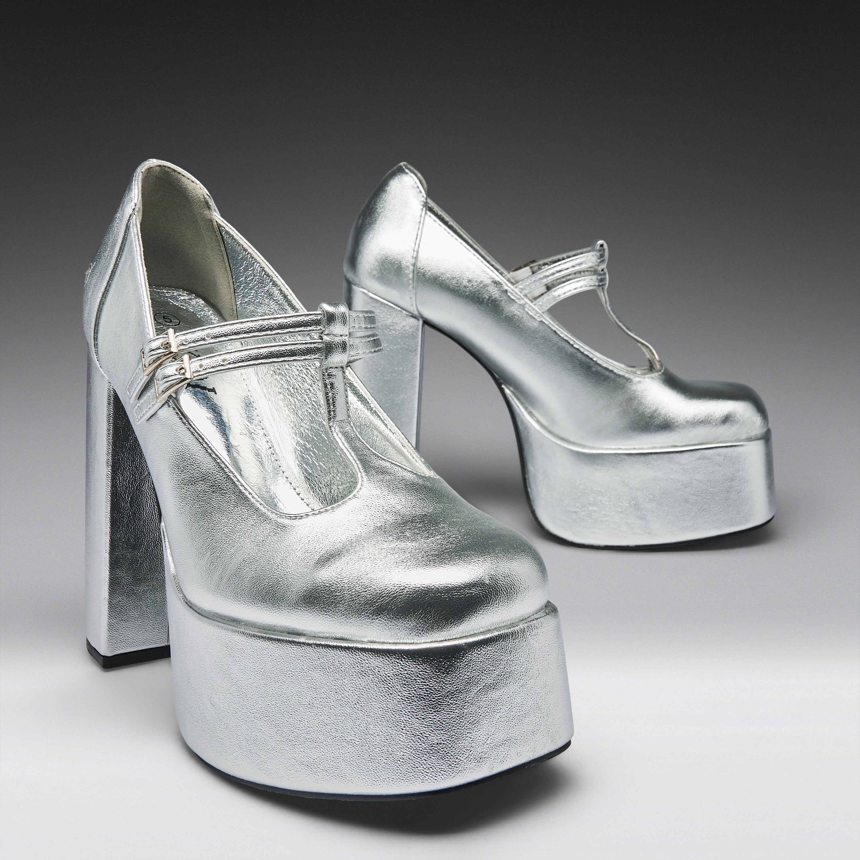 A pair of metallic silver platform Mary Jane high heels with block heels and ankle straps, photographed on a dark-to-light gray gradient studio background.