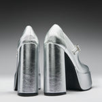 A pair of metallic silver high-heeled pumps with block heels and platform soles, shown from the back on a dark-to-light gray gradient studio background.