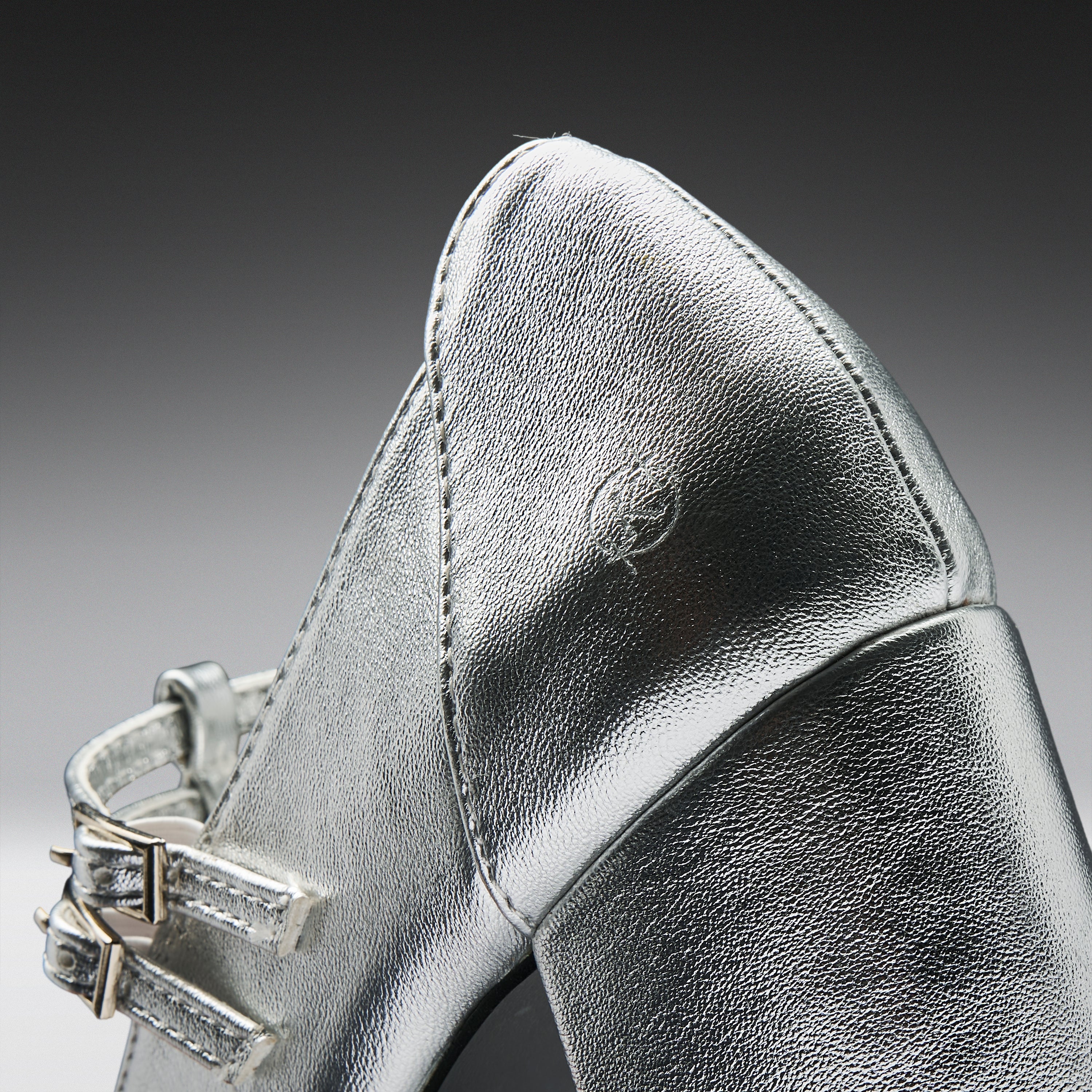 Close-up of a metallic silver leather shoe with a pointed toe and double strap detailing on a dark gray gradient background.