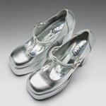 A pair of metallic silver Mary Jane platform heels with block heels and buckle straps, displayed on a neutral gray gradient background. The brand name 'KOT' is visible on the insoles.