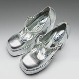 A pair of metallic silver Mary Jane platform heels with block heels and buckle straps, displayed on a neutral gray gradient background. The brand name 'KOT' is visible on the insoles.