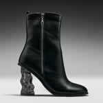 A black leather ankle boot with a sculptural, wavy transparent block heel, shown from a side profile against a dark gray studio background.