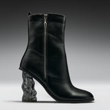 A black leather ankle boot with a sculptural, wavy transparent block heel, shown from a side profile against a dark gray studio background.