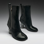 A pair of black leather ankle boots with a distinctive split toe and sculptural clear resin animal-head heels, shown against a dark gray studio background.