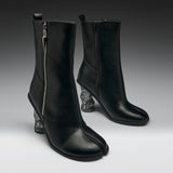 A pair of black leather ankle boots with a distinctive split toe and sculptural clear resin animal-head heels, shown against a dark gray studio background.