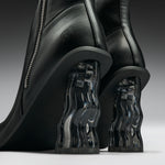 Close-up of a pair of black leather ankle boots with sculptural, wavy transparent acrylic block heels on a dark gray studio background.