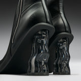 Close-up of a pair of black leather ankle boots with sculptural, wavy transparent acrylic block heels on a dark gray studio background.