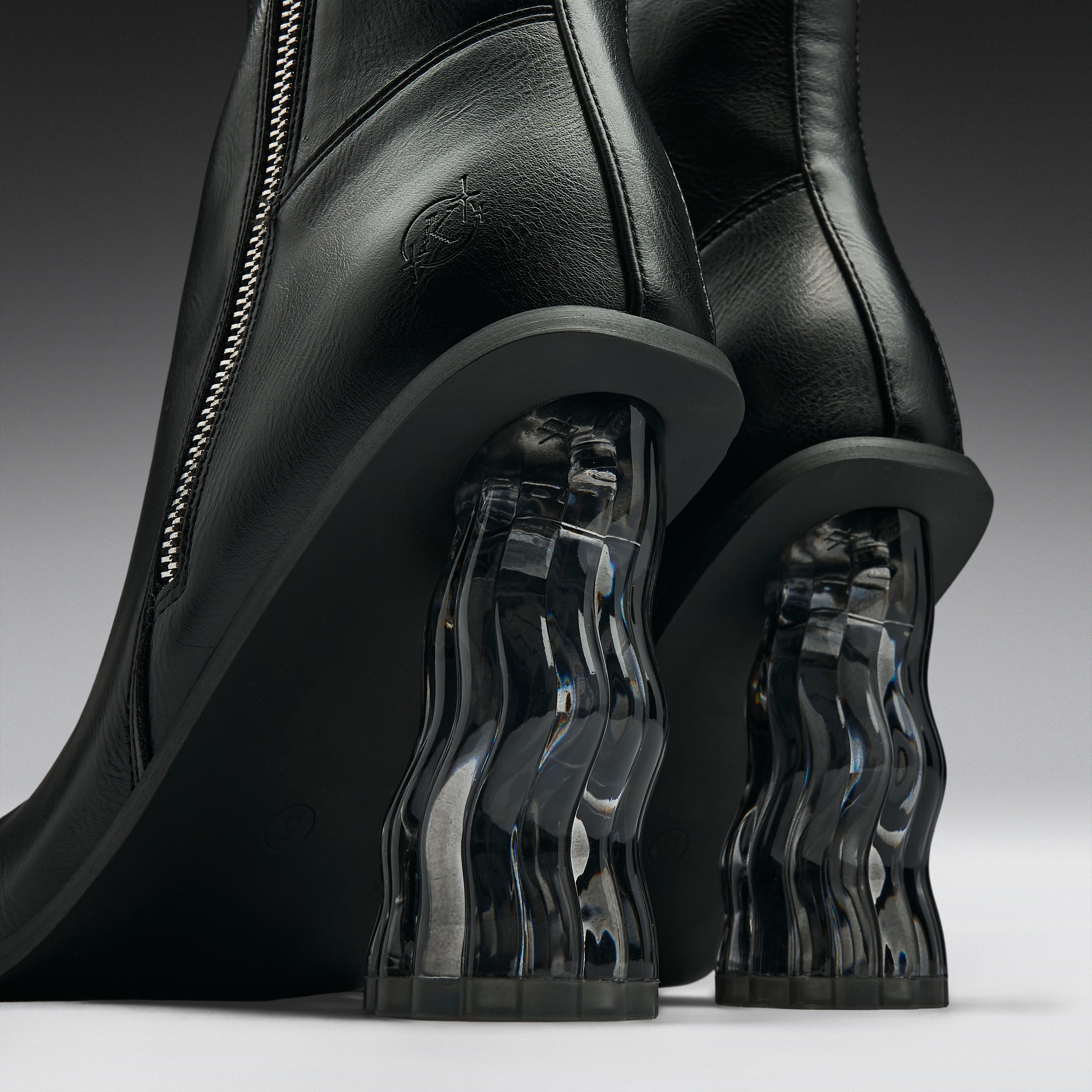 Close-up of a pair of black leather ankle boots with sculptural, wavy transparent acrylic block heels on a dark gray studio background.