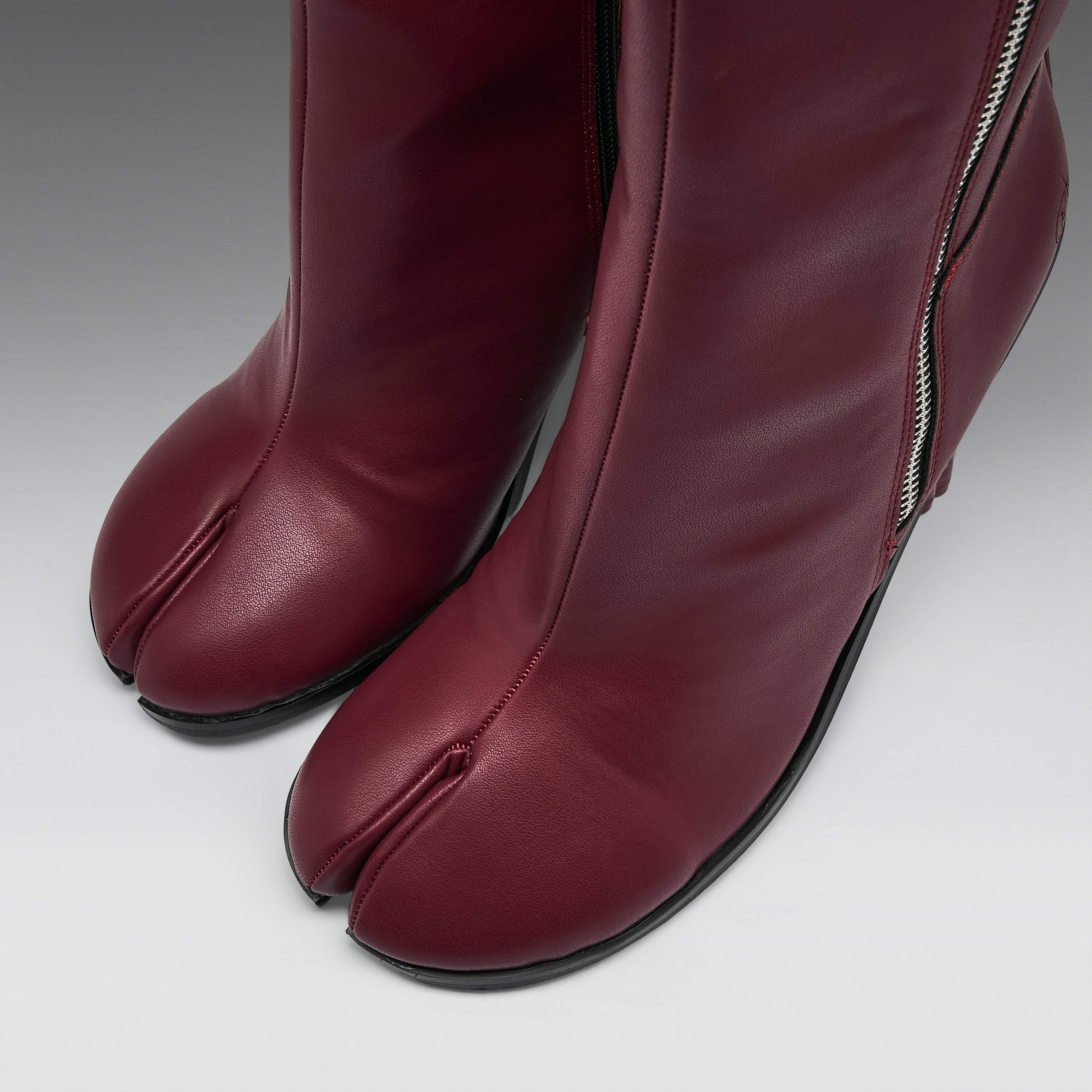 Close-up of a pair of burgundy leather tabi-style boots with a side zipper, shown on a neutral gray background.