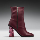 A single women's ankle boot in burgundy leather with a silver side zipper, a chunky wavy transparent pink block heel, and a black outsole, shown on a dark-to-light gray gradient studio background.