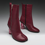 A pair of dark burgundy leather ankle boots with a split toe, side zippers, and chunky translucent pink block heels, shown on a gray gradient studio background.