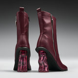 A pair of burgundy leather ankle boots with chunky black and translucent pink wavy block heels, shown from the back and side on a gray gradient background.