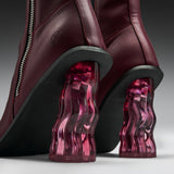 Close-up of a pair of burgundy leather ankle boots with sculptural, wavy translucent magenta-pink block heels and black soles.