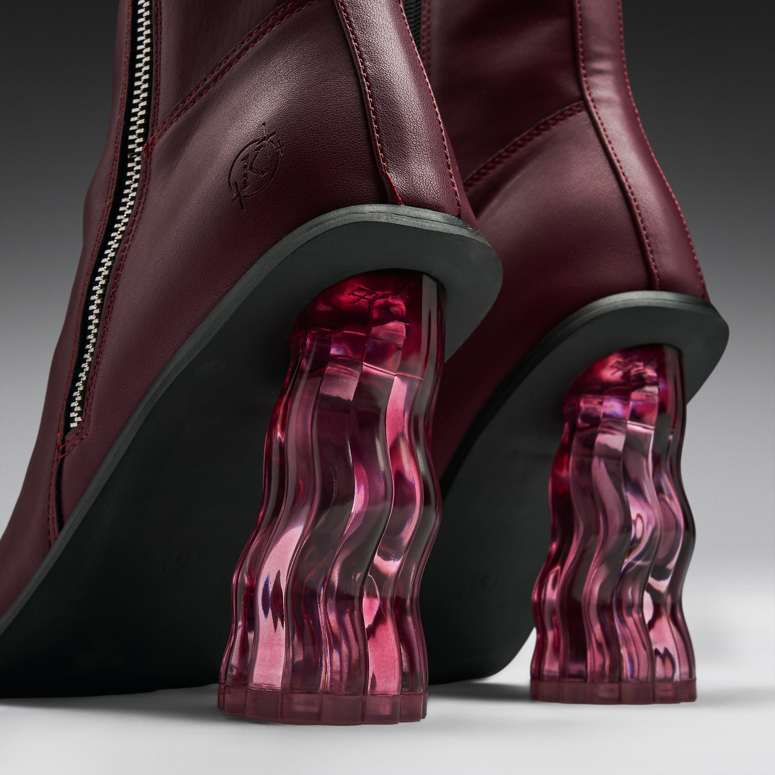 Close-up of a pair of burgundy leather ankle boots with sculptural, wavy translucent magenta-pink block heels and black soles.