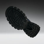 A close-up of a black rubber shoe sole with deep tread patterns and embossed logo details, shown against a dark-to-light gray gradient background.