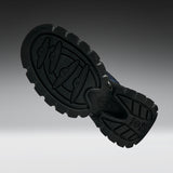 A close-up of a black rubber shoe sole with deep tread patterns and embossed logo details, shown against a dark-to-light gray gradient background.