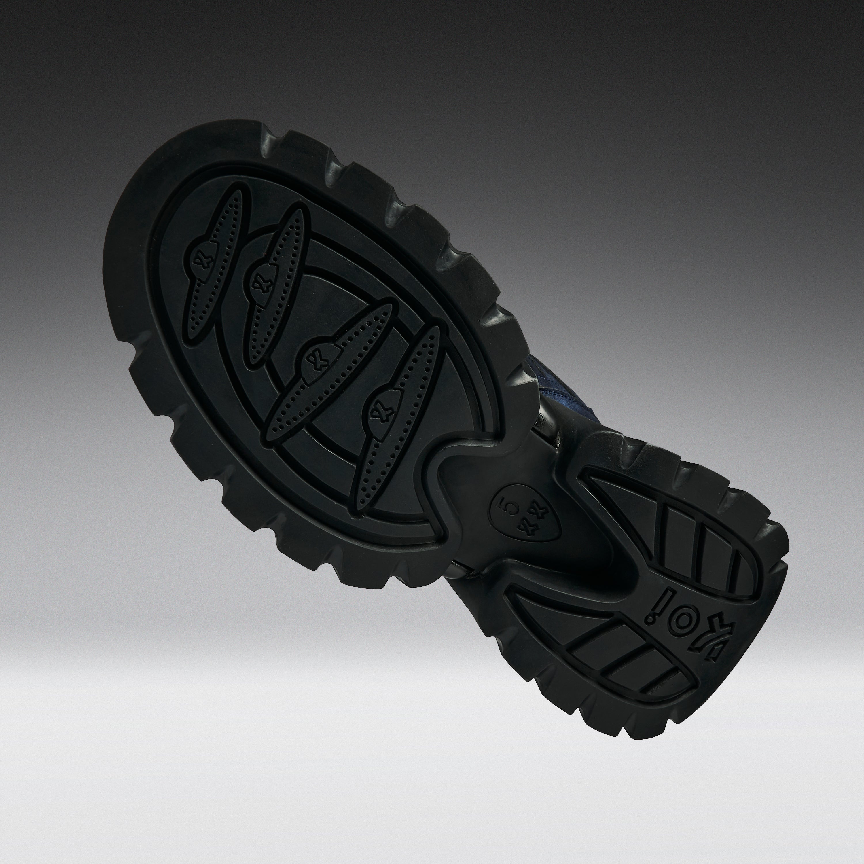 A close-up of a black rubber shoe sole with deep tread patterns and embossed logo details, shown against a dark-to-light gray gradient background.