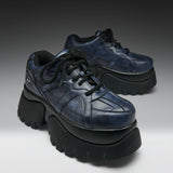 A pair of chunky low-top platform sneakers in distressed navy blue leather with thick black platform soles and black laces, photographed on a dark-to-light gray gradient studio background.