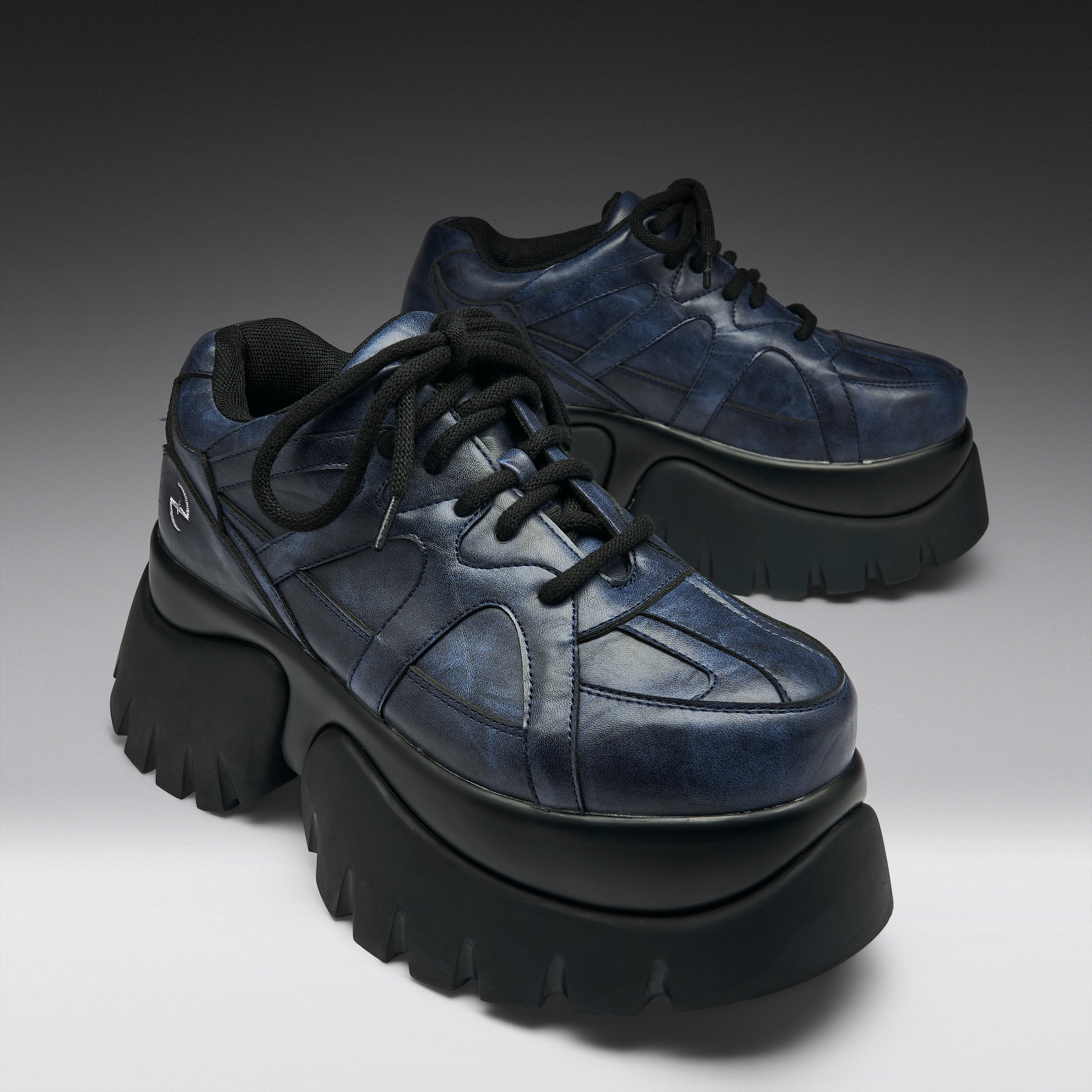 A pair of chunky low-top platform sneakers in distressed navy blue leather with thick black platform soles and black laces, photographed on a dark-to-light gray gradient studio background.
