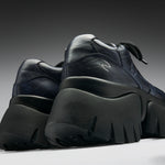 A pair of chunky low-top sneakers in dark navy blue leather with thick black platform soles, shown from a low angle against a dark gray gradient background.