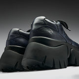 A pair of chunky low-top sneakers in dark navy blue leather with thick black platform soles, shown from a low angle against a dark gray gradient background.