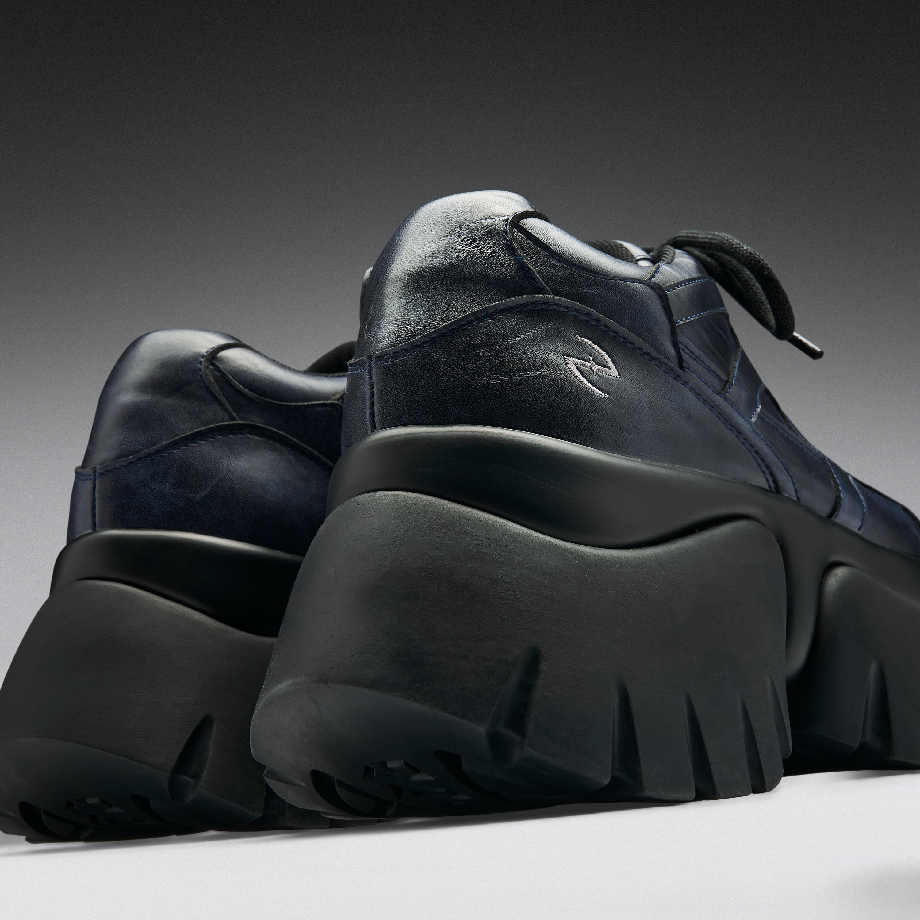 A pair of chunky low-top sneakers in dark navy blue leather with thick black platform soles, shown from a low angle against a dark gray gradient background.