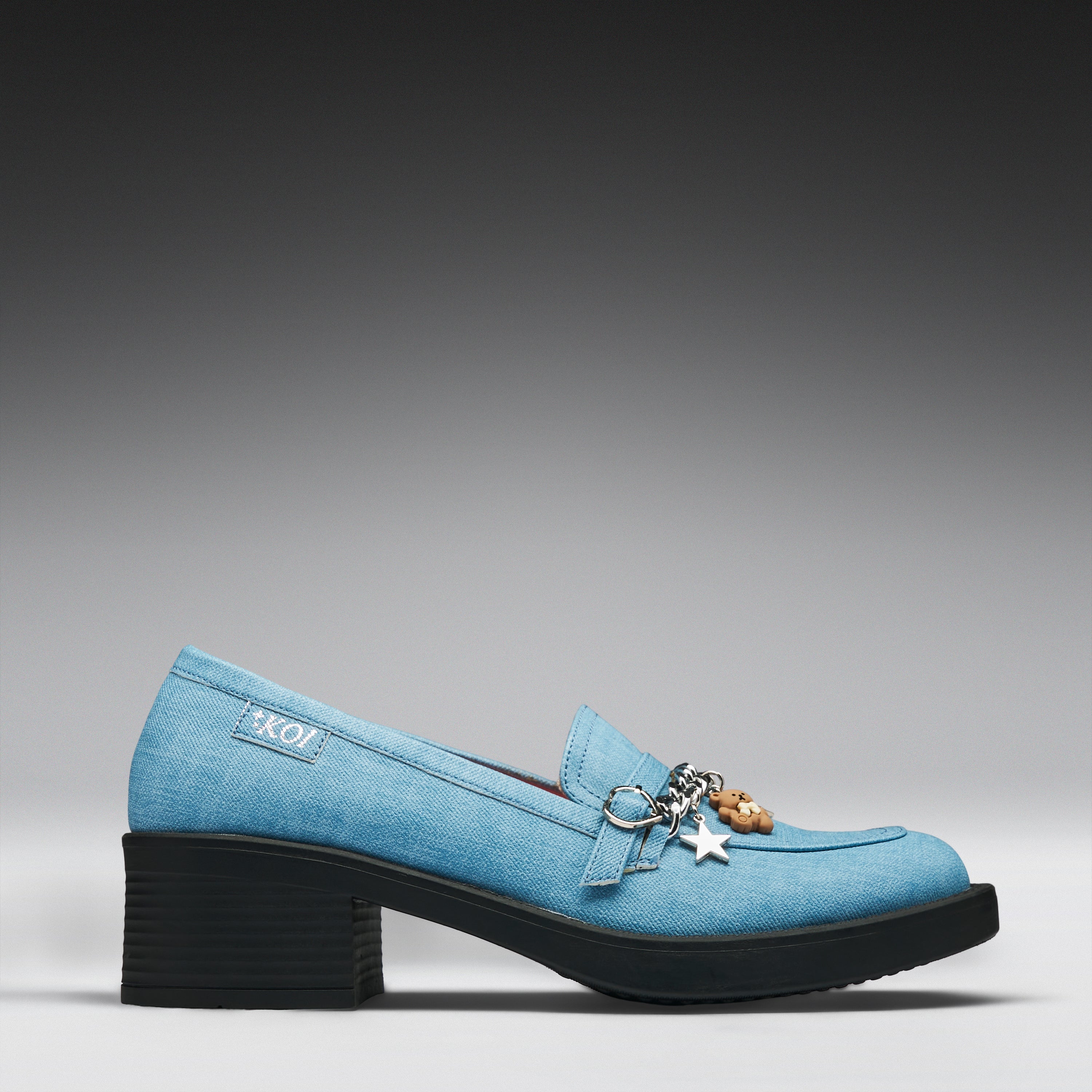 A light blue denim-textured loafer-style shoe with a chunky black block heel, decorative metal rings, and hanging charms, shown in side profile on a gray gradient background.