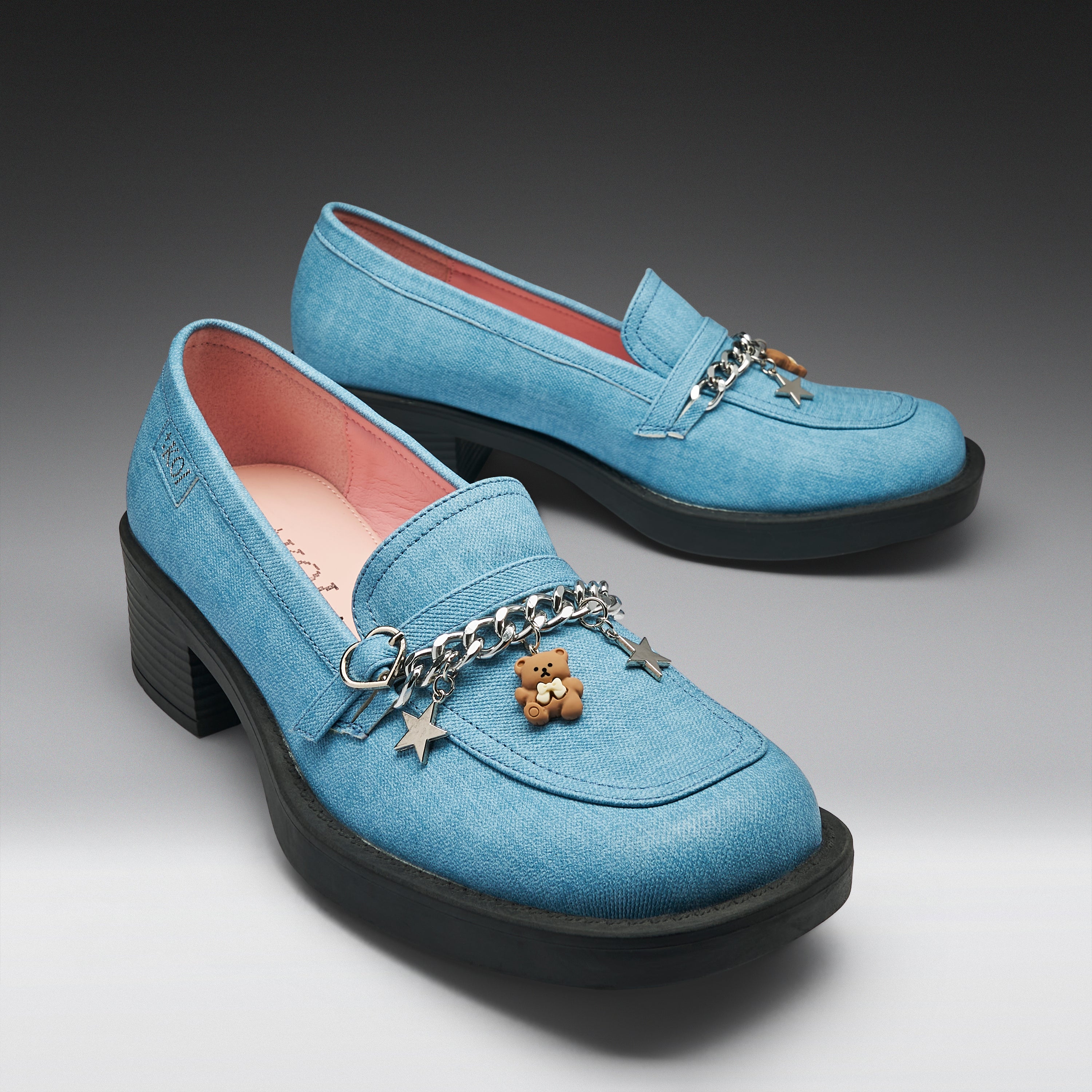 A pair of light blue suede loafers with chunky black platform heels, decorated with a silver chain, star charms, and a small teddy bear charm. The shoes are photographed on a dark gray to light gray studio background.