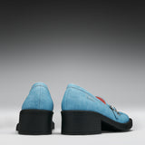 A pair of light blue denim loafers with chunky black block heels, shown from the back against a dark-to-light gray gradient studio background.