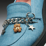 A close-up of a light blue denim shoe decorated with a silver chain accessory featuring a small brown bear charm and two silver star charms.