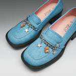 A pair of light blue denim-like loafers with black soles, decorated with a silver chain, star charms, and a small teddy bear charm. The insoles are pink with the brand name 'IOKI' printed on them.