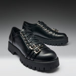 A pair of black leather lace-up shoes with chunky lug soles and decorative gold-tone buckle straps on the sides, photographed on a dark-to-light gray gradient studio background.