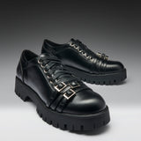 A pair of black leather lace-up shoes with chunky lug soles and decorative gold-tone buckle straps on the sides, photographed on a dark-to-light gray gradient studio background.