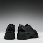 A pair of black leather lace-up shoes with chunky lug soles, shown from the back and side against a dark gray gradient background.