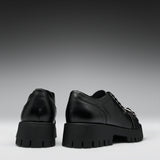 A pair of black leather lace-up shoes with chunky lug soles, shown from the back and side against a dark gray gradient background.