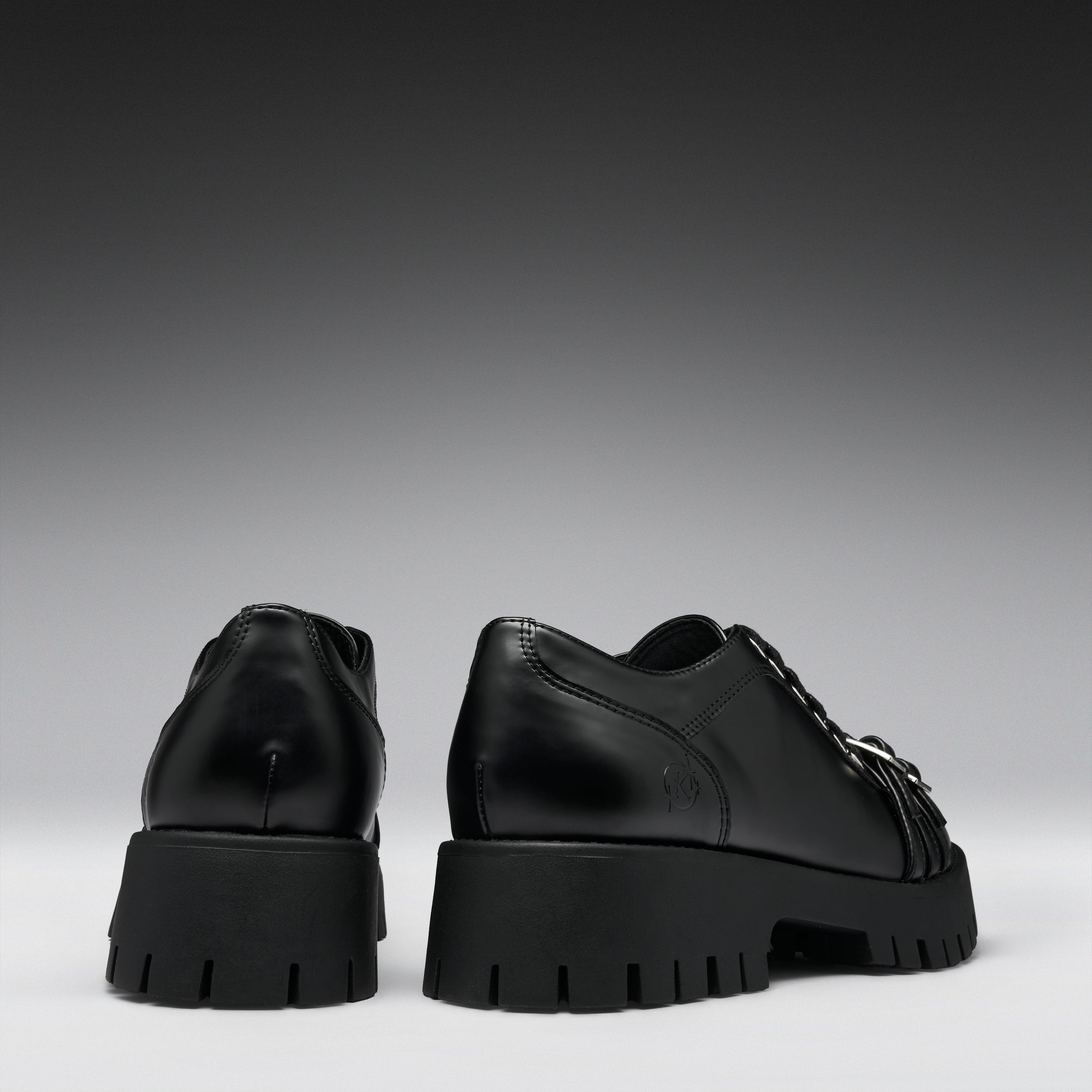A pair of black leather lace-up shoes with chunky lug soles, shown from the back and side against a dark gray gradient background.