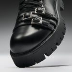 Close-up of a black leather lace-up boot with two silver buckle straps and a chunky lug sole, photographed on a gray studio background.